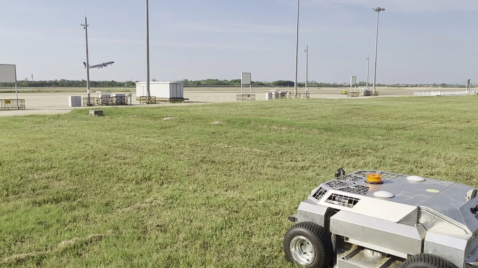 Amotrol A60i autonomous mower cutting grass on an airport apron with an aircraft climbing out in the background, light poles and ground equipment visible