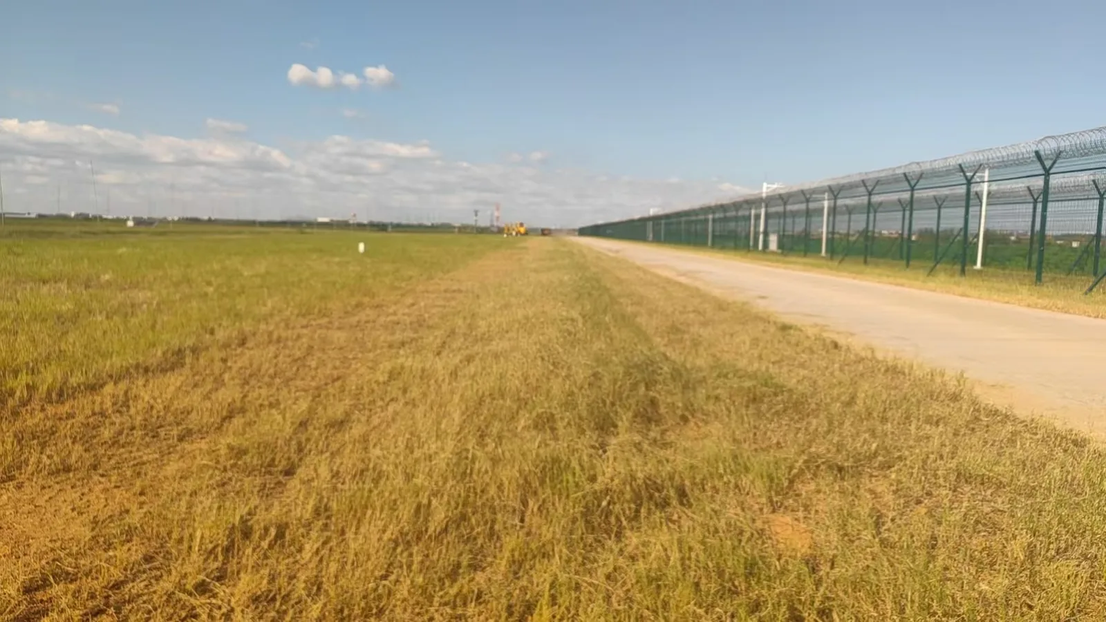 Airport perimeter fence line with a sharp cut line between tall uncut grass and freshly mowed grass, security fence and patrol road on the right